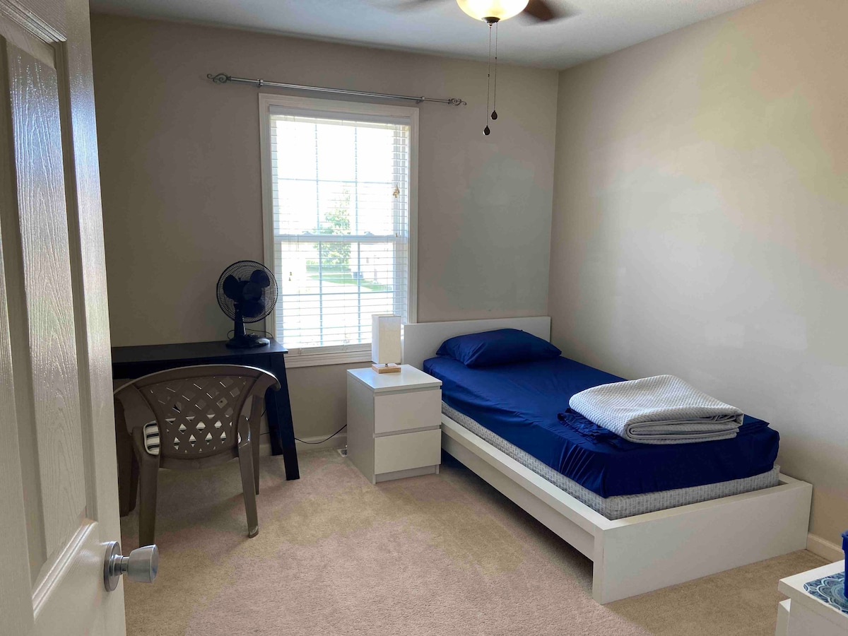 A private room features a spacious bed with blue bedding and a light blanket, accompanied by a small nightstand. A desk with a chair is positioned near a window, which allows natural light to fill the room. A standing fan is visible in the corner.