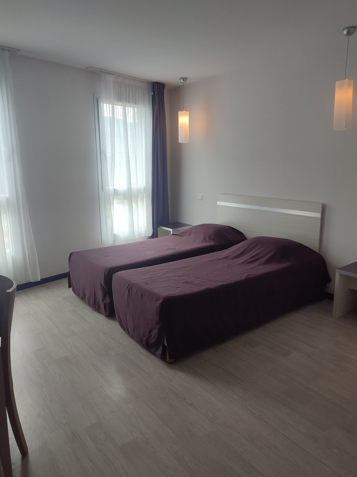 A bright and simple room features two twin beds, each covered with deep purple bedspreads. Natural light enters through large windows with sheer curtains, illuminating the light wood flooring. Soft lighting fixtures hang from the ceiling, adding to the uncluttered ambiance.