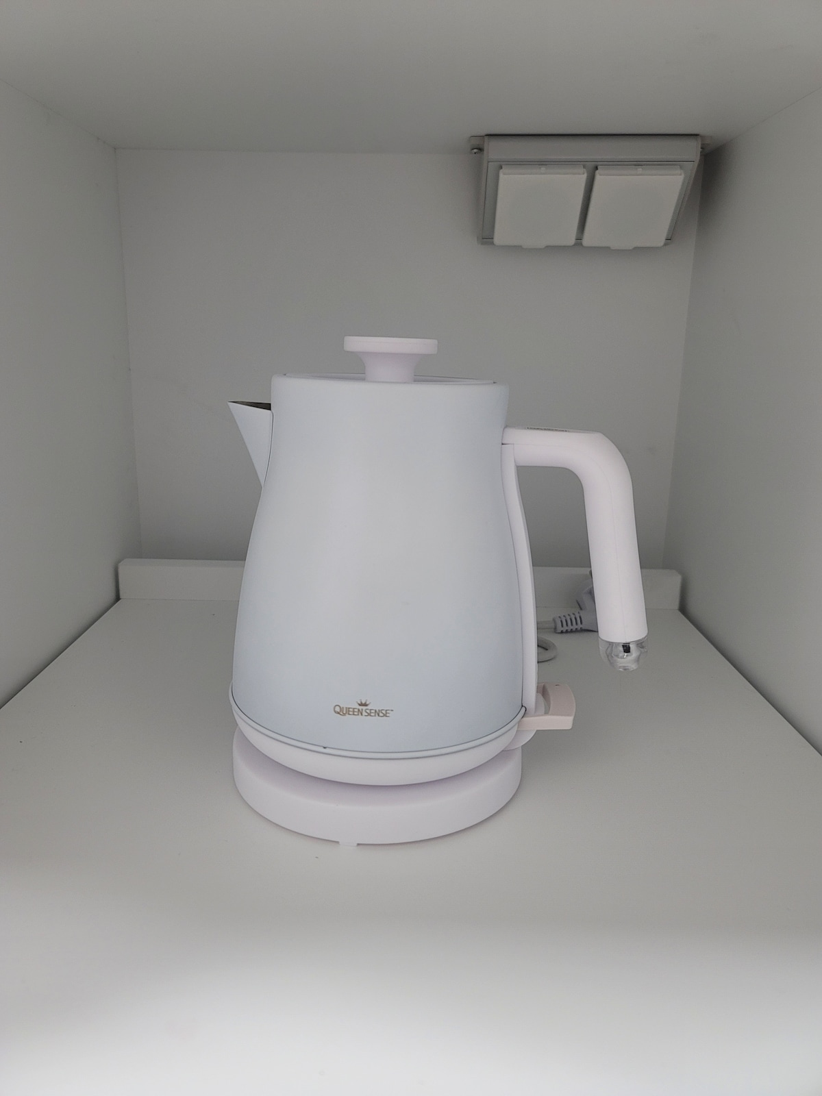 An electric kettle sits on a white shelf, featuring a sleek design with a smooth finish. The kettle is equipped with a handle and spout for easy pouring. The surrounding area is minimalistic, with electrical outlets visible in the background.