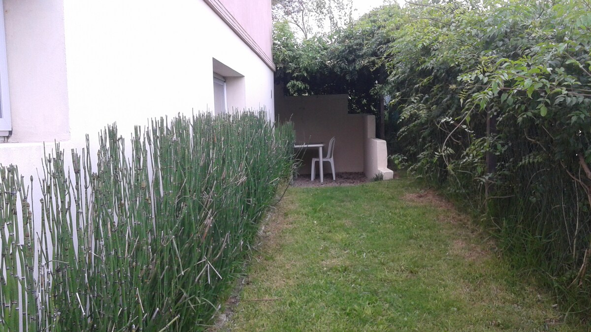 A small garden area is bordered by green foliage, creating a private outdoor space. A table and chair set are visible, providing a serene spot for relaxation. The lawn is well-kept, enhancing the tranquil atmosphere of the corner garden.