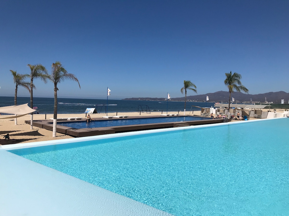An expansive infinity pool mirrors the clear blue sky, leading the eye toward the horizon of the ocean. Palm trees sway gently beside the pool, while beach loungers and umbrellas are positioned along the sandy shore, creating a relaxed outdoor atmosphere.