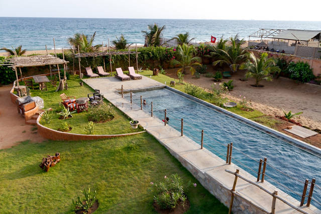 A tropical garden features a long, serene pool bordered by lush greenery. Sun loungers are positioned nearby, with shaded seating arrangements visible. In the background, the ocean waves create a scenic backdrop, completing the tranquil atmosphere of the outdoor space.