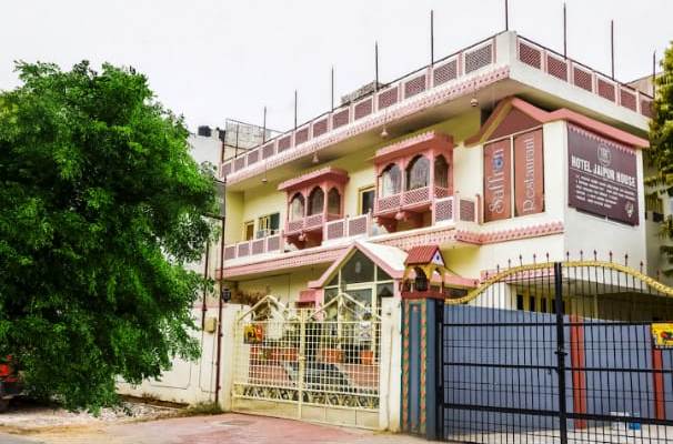 A multi-storied building reflects traditional architecture with decorative elements on the upper level. A gated entrance provides access to the property, flanked by greenery. The exterior walls are painted in soft tones, and large windows enhance natural light in the spaces.