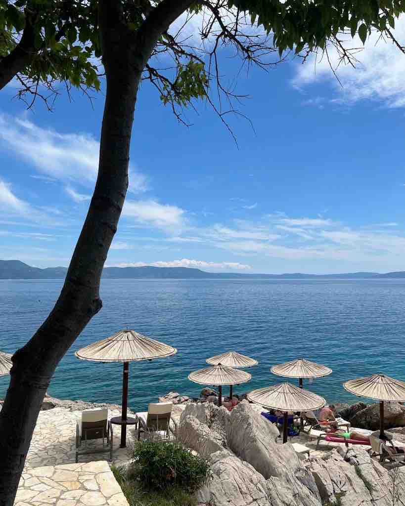 A serene coastal view features several wooden umbrellas shading lounge chairs positioned on rocky terrain. The calm blue sea extends toward a distant coastline, framed by lush green trees and a clear sky with scattered clouds.
