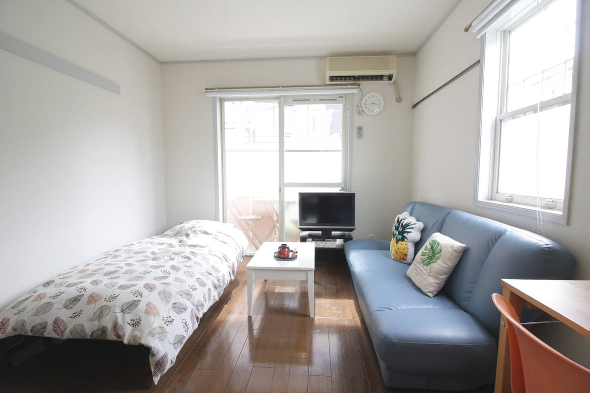 A bright and functional living space is displayed, featuring a bed with a patterned duvet cover, a blue sofa, and a small coffee table. A television sits on a stand near the window, which is dressed with light curtains, allowing natural light to fill the room.