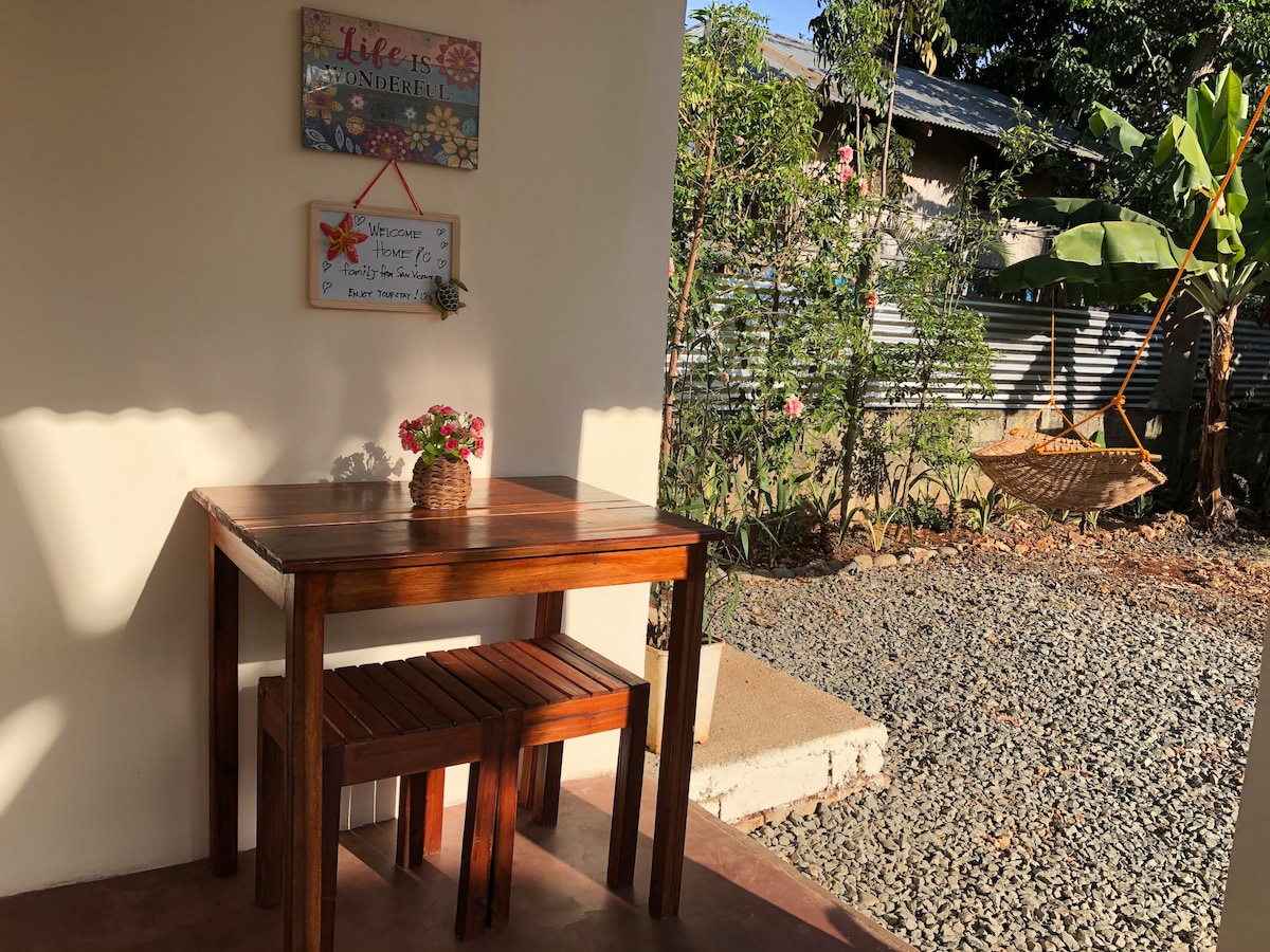A small outdoor seating area features a wooden table accompanied by two matching stools. A decorative wall sign with floral artwork welcomes visitors. Lush greenery and a hanging chair create a tranquil atmosphere, complemented by gravel flooring and natural light illuminating the space.