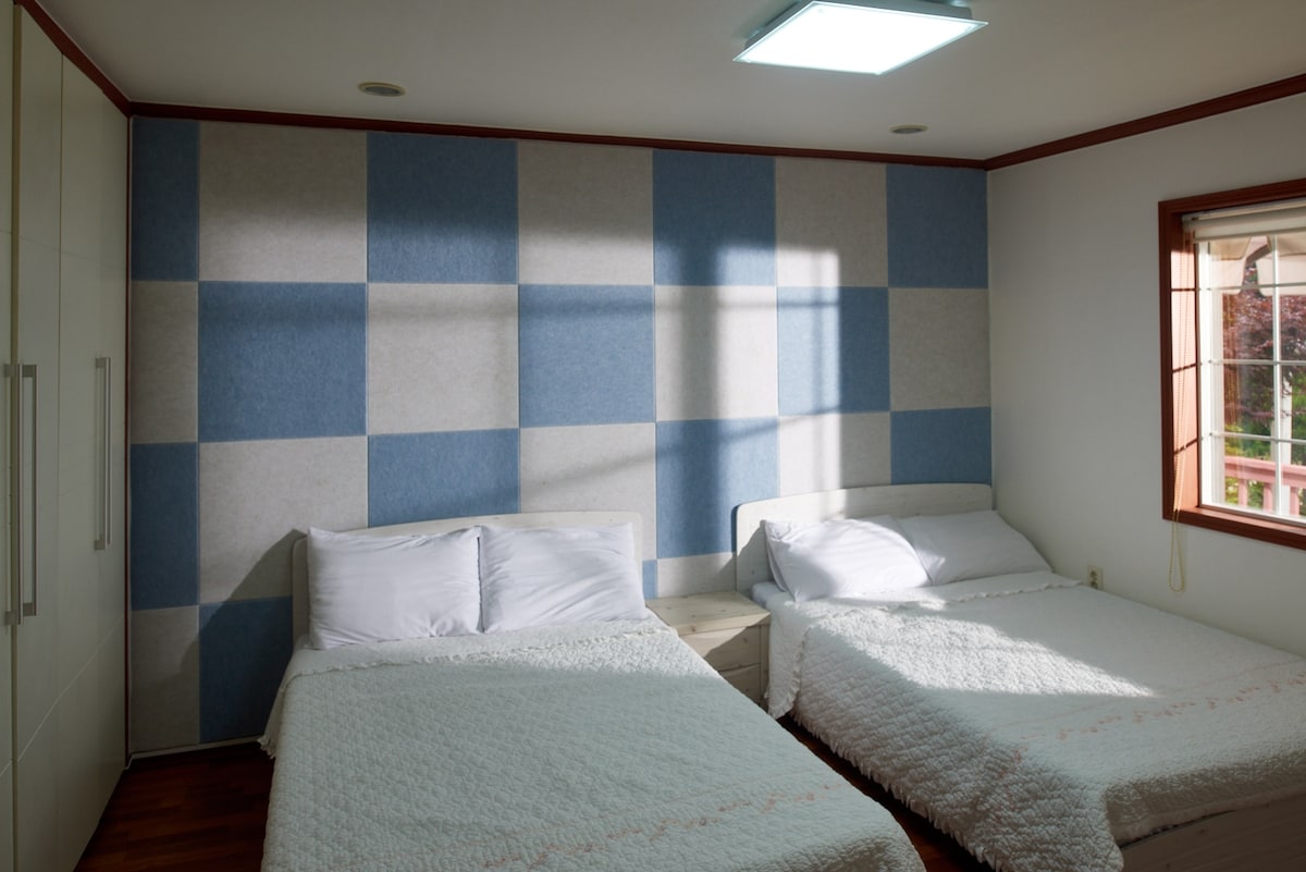 A well-lit bedroom features two spacious beds with white bedding, complemented by a large window allowing natural light to brighten the room. The soft blue and gray checkered wall creates a calm atmosphere, while the wooden floor adds warmth to the space.