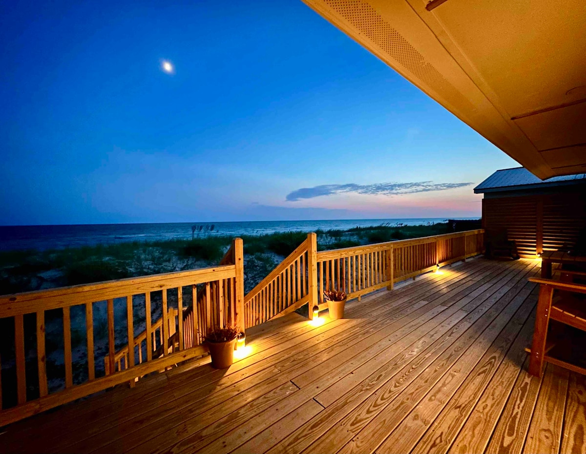 A spacious deck is illuminated by softly glowing lights, offering expansive views of the ocean and sandy shoreline at dusk. The sky transitions from blue to soft lavender, with the moon visible above. Wooden railings frame the deck, enhancing the outdoor space.