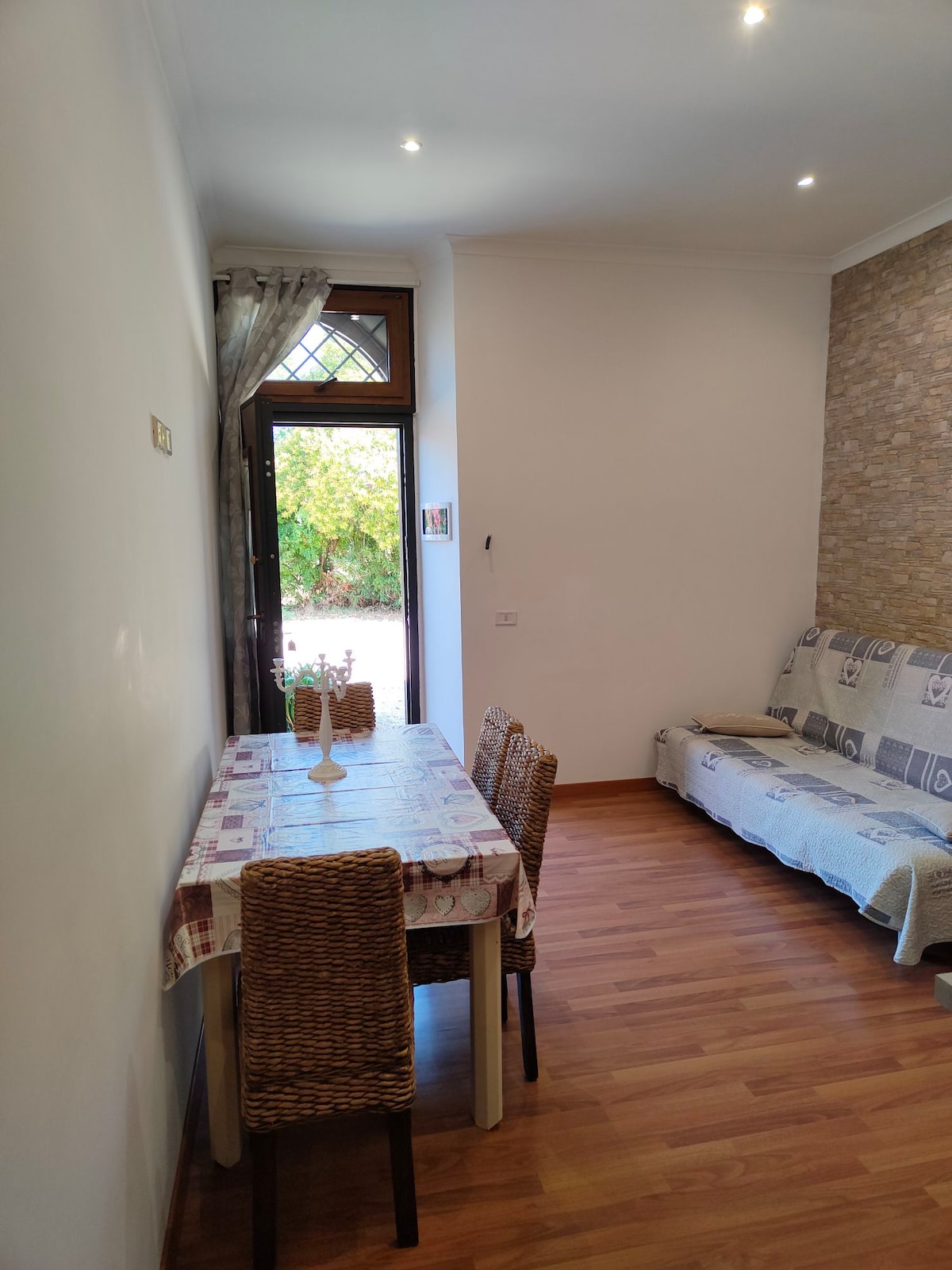 A bright dining area features a wooden table set for four, surrounded by woven chairs. Natural light enters through a large door that opens to a garden view. A cozy sofa is positioned against the wall, enhancing the inviting simplicity of the space.