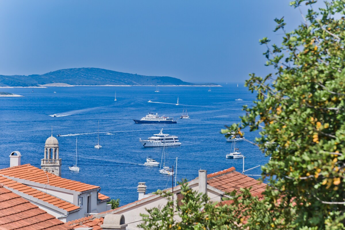 A panoramic view showcases the sparkling blue waters of the sea dotted with various boats. The horizon features distant islands under a clear sky, while the foreground includes terracotta rooftops and green foliage, providing a serene backdrop.
