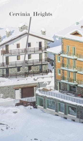A snow-covered landscape showcases two distinct buildings, with a prominent sign reading 'Cervinia Heights.' The structure on the left features wooden balconies and stone accents, while the building on the right presents a modern design with expansive glass windows. Snow blankets the surrounding area.