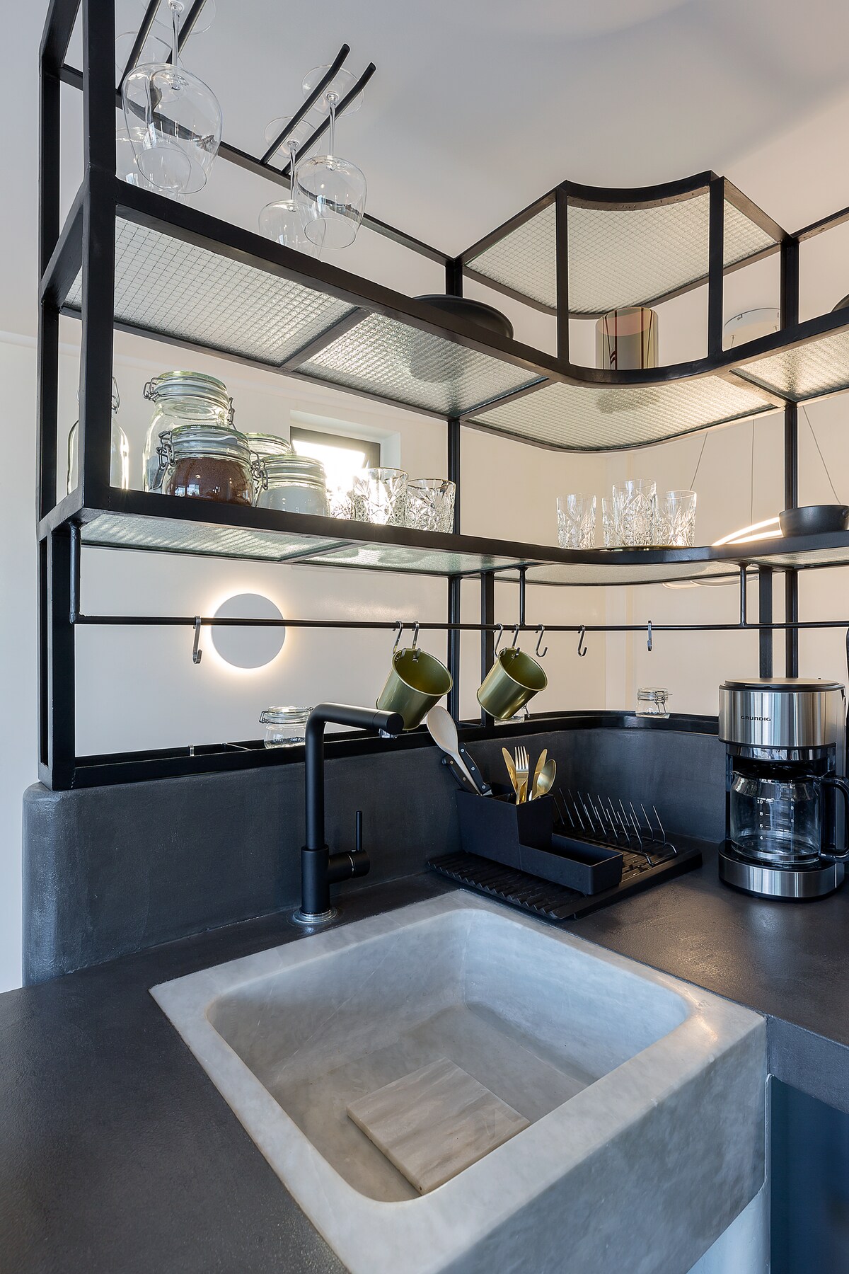 The kitchen showcases a modern design with black metal shelving, displaying glassware and jars. A concrete sink is positioned below a sleek faucet. A coffee maker and dish rack are neatly arranged on the counter, enhancing the space's functionality.