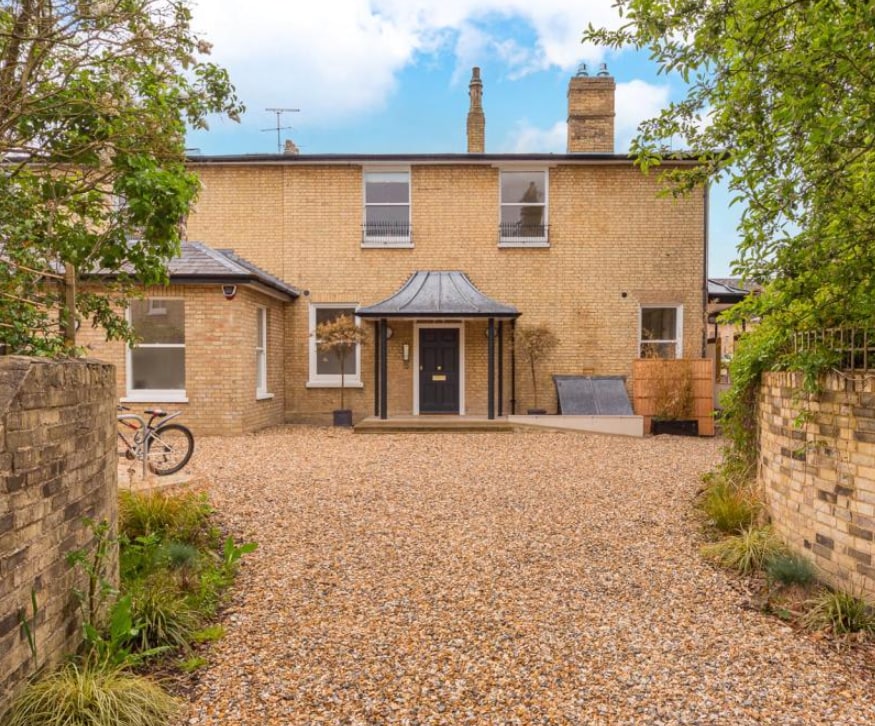 The exterior of a historic villa is showcased, featuring a gravel driveway that leads to a welcoming entrance. The two-story building has a blend of brick and modern design, with large windows and a distinctive roof creating an inviting atmosphere. Lush greenery frames the property.