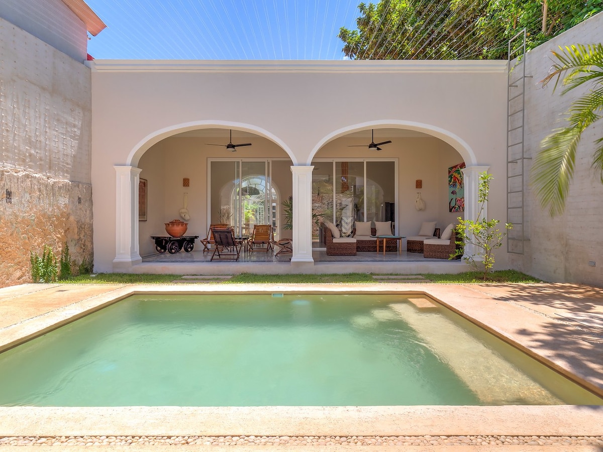A charming outdoor space features a rectangular swimming pool surrounded by a grassy area. Arched doorways lead into a living area furnished with comfortable seating arrangements and decorative elements. Two ceiling fans enhance the open-air design, complemented by lush greenery in the background.