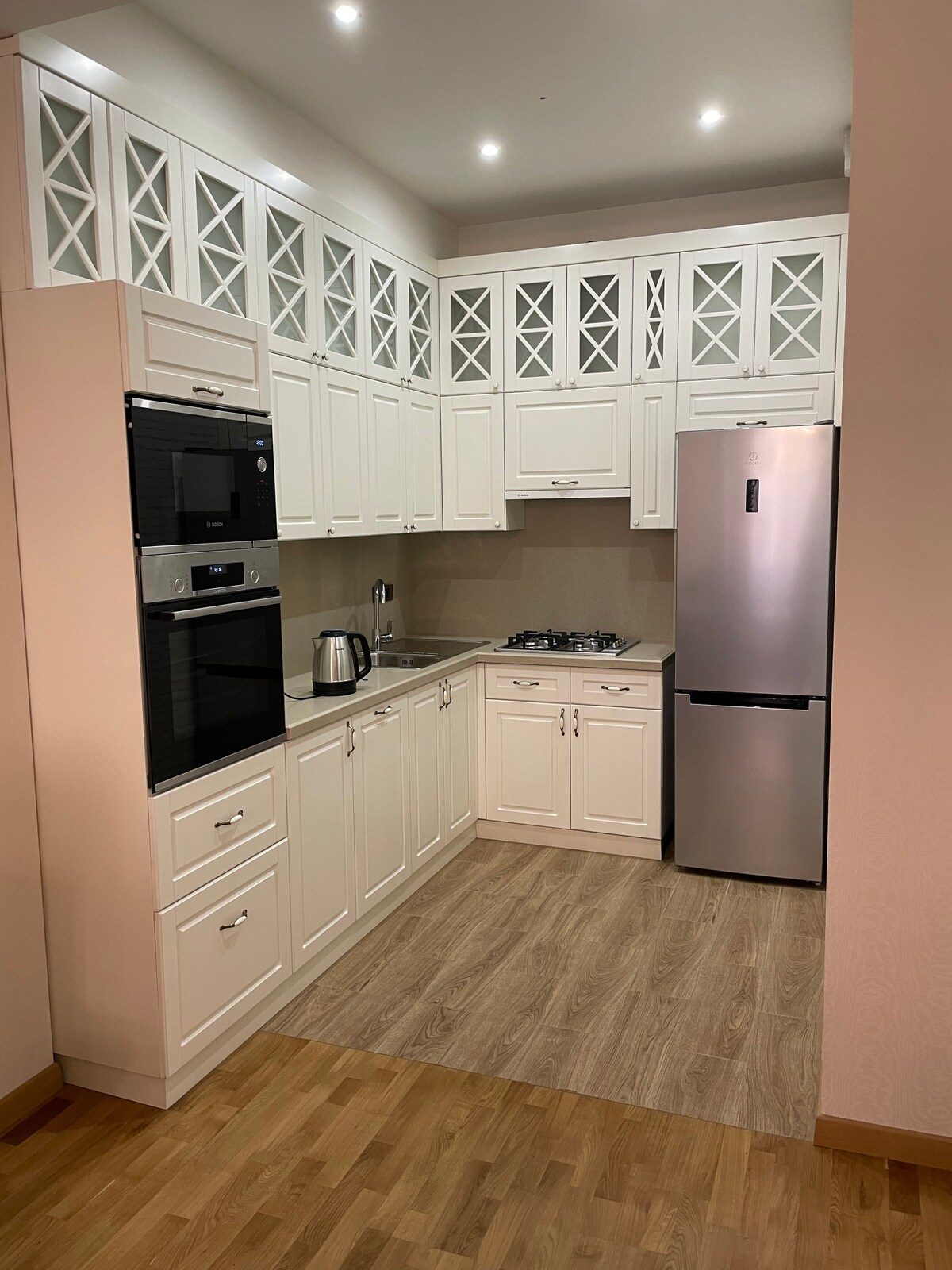 The kitchenette showcases sleek white cabinetry with glass-panel accents, providing a bright and airy feel. Modern appliances, including an oven and refrigerator, are featured alongside a stovetop. The flooring consists of light wood, creating a warm contrast with the design.