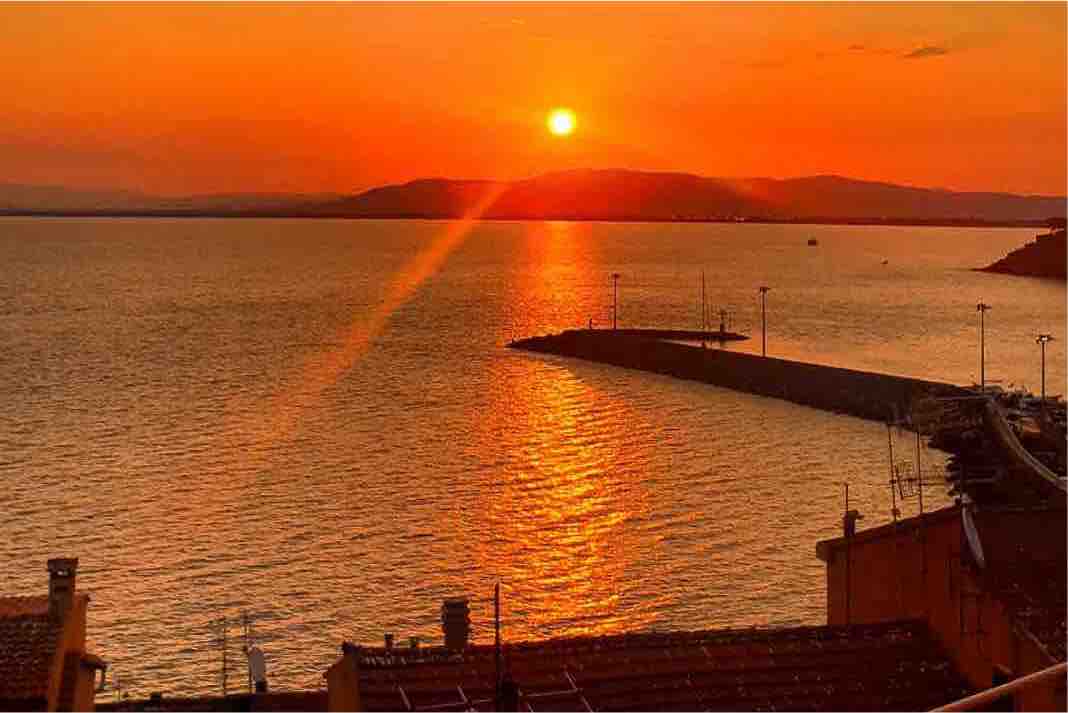 A tranquil sunset casts an orange glow over the water, reflecting vibrant hues across the surface. The silhouette of distant hills can be seen on the horizon, while a pier extends into the sea, creating a sense of depth and openness.