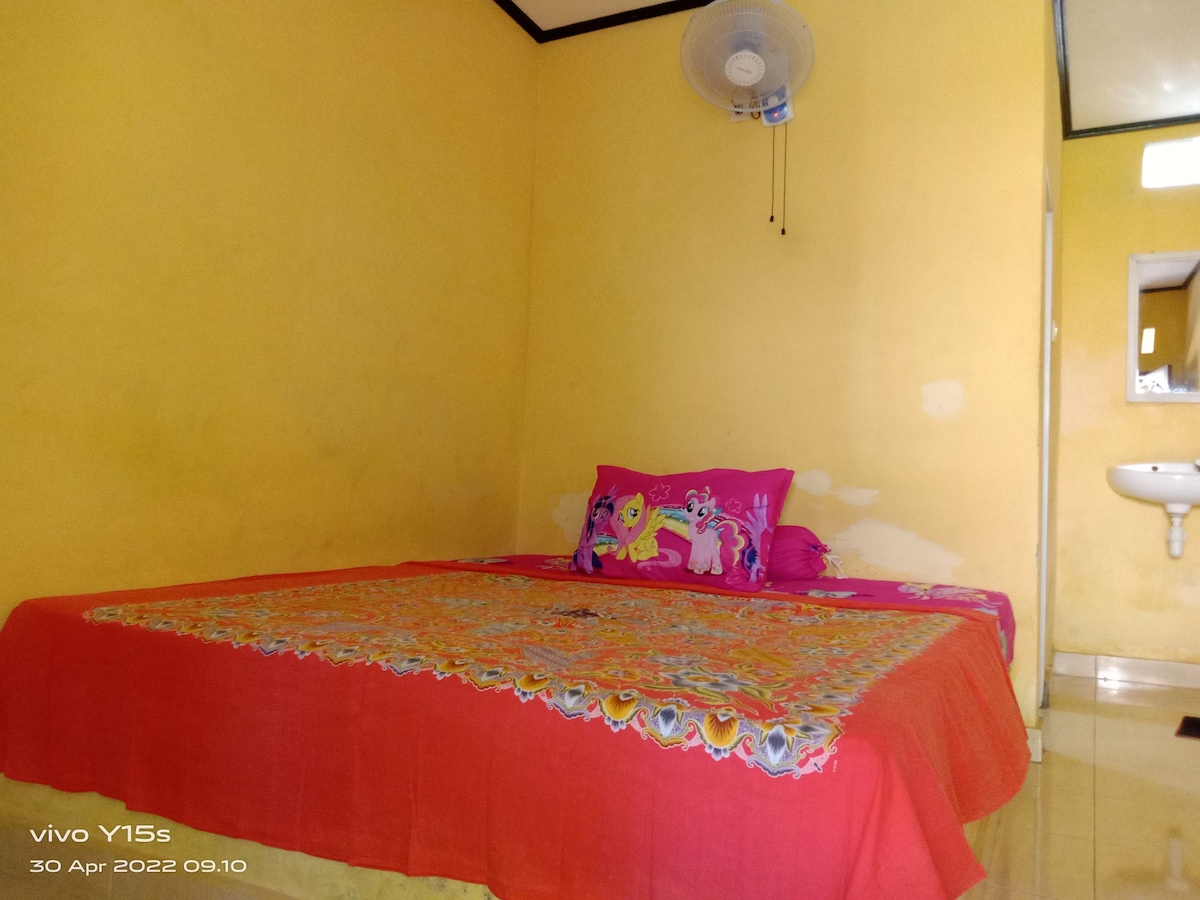 A spacious sleeping area features bright yellow walls and a colorful bedspread adorned with floral patterns. A large bed is placed centrally, accompanied by decorative pillows. A ceiling fan is mounted above, and a small sink with a mirror is visible in the background.