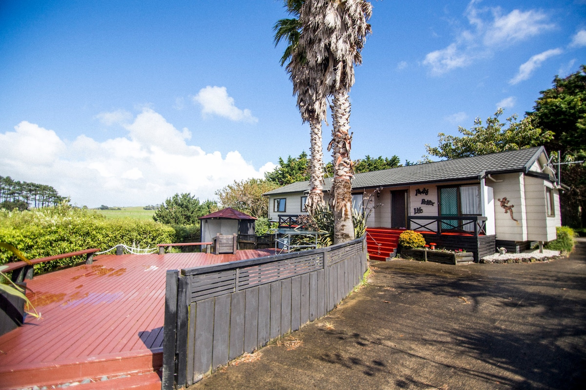A rural home is set amidst lush greenery, featuring a spacious deck area with outdoor seating. Tall palm trees frame the entrance, leading to a welcoming façade with light-colored siding and a dark roof. The clear blue sky enhances the tranquil setting.