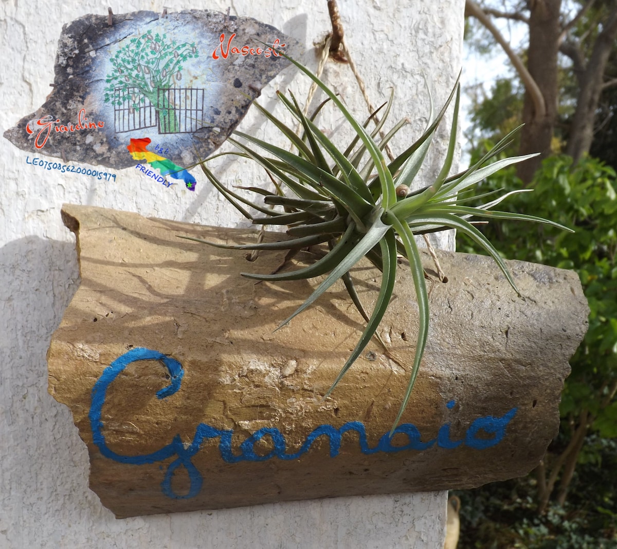 A rustic sign displaying the word 'Granaio' is attached to a textured wall. The sign is crafted from natural materials, and an air plant is affixed above it, adding a touch of greenery. The surrounding garden is softly blurred in the background.