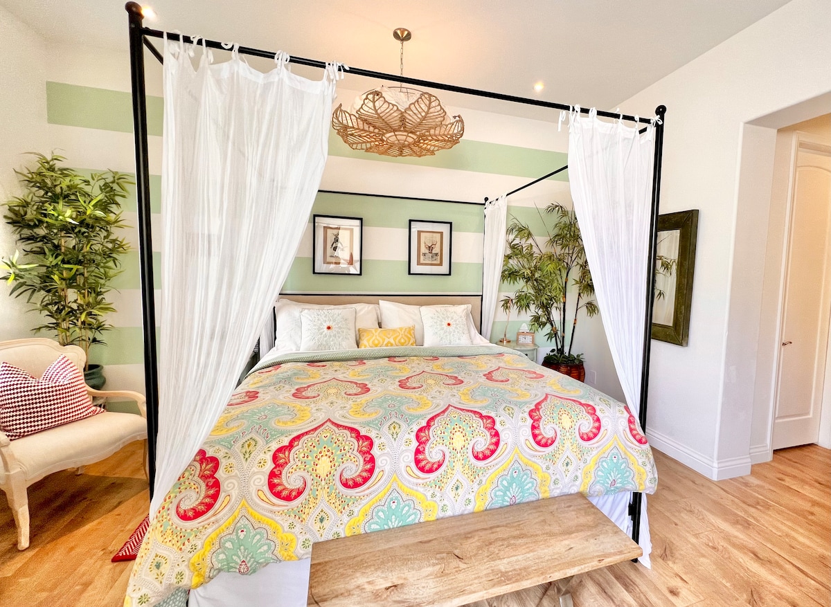 A spacious bedroom features a four-poster bed draped with sheer white curtains. The bed is adorned with a vibrant patterned comforter. Flanking the bed are potted plants and framed artwork on the walls, with an inviting seating area visible in the corner.
