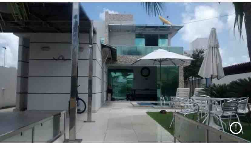 The image showcases a modern two-story house with large glass panels, allowing natural light to fill the space. A private pool is visible, surrounded by lounge chairs and outdoor seating. Sun umbrellas provide shade, while lush greenery adds a touch of nature.