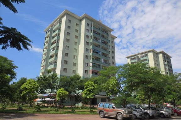 A cluster of residential buildings stands amongst greenery, featuring multiple floors and balconies. A clear blue sky is visible above, while parked cars line the foreground and trees add natural beauty to the area.