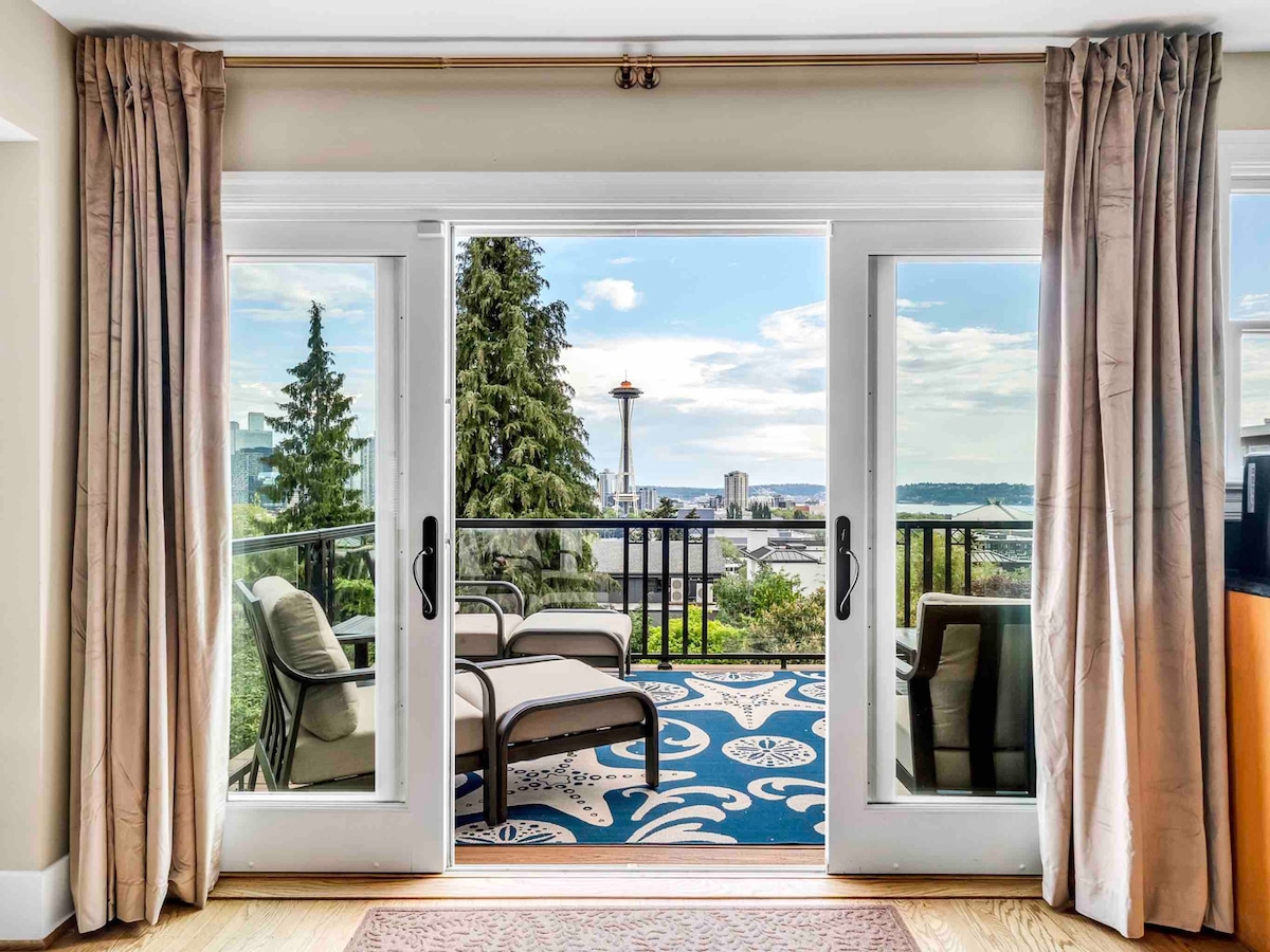 Sliding glass doors open onto a private balcony featuring comfortable outdoor seating. The view reveals the Space Needle and surrounding greenery, with the cityscape and bay visible in the background. Sheer curtains frame the entrance, adding a touch of elegance to the space.
