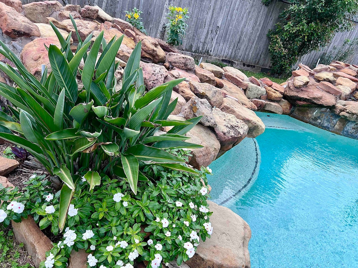 A tranquil pool area is surrounded by natural stone landscaping and lush greenery. Tropical plants and white flowers complement the vibrant blue water. The setting offers a serene atmosphere, perfect for relaxation under the sun.