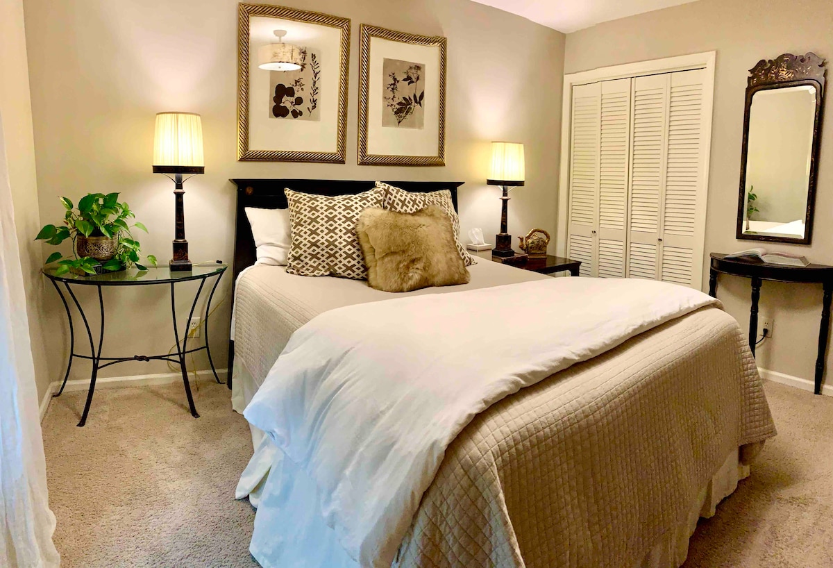 A cozy bedroom contains a spacious bed dressed in soft linens and decorative pillows. Two bedside lamps provide gentle illumination. Wall art is displayed above the bed, and a plant adds a touch of greenery. A closet and a small dressing table are visible, enhancing functionality.
