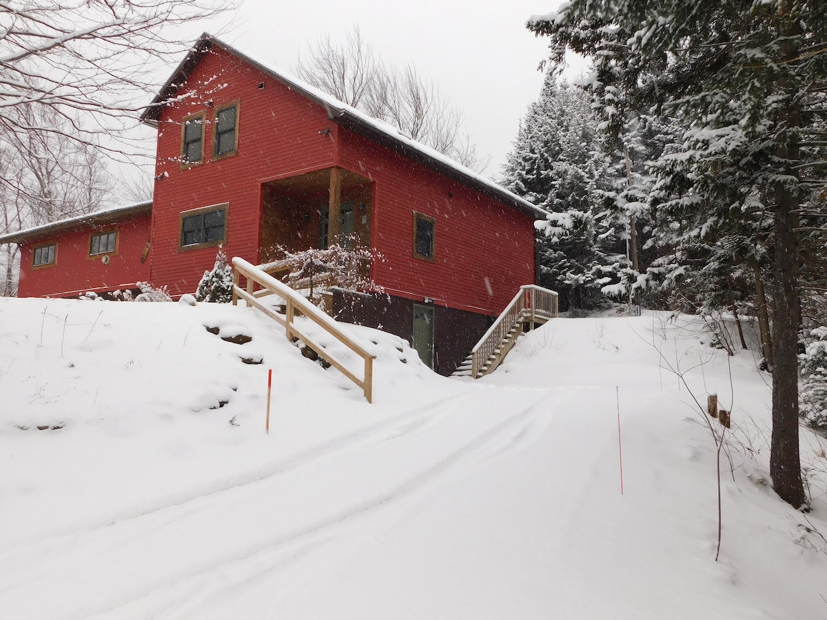 A red two-story house is situated among snow-covered trees, with a gently sloping driveway leading up to the entrance. Soft snowfall creates a serene winter atmosphere, while the surrounding natural landscape is blanketed in white.