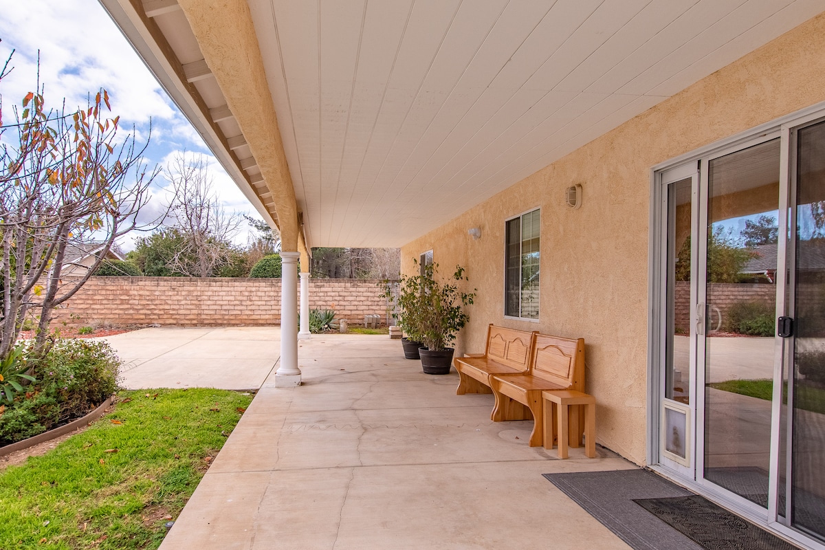 A covered outdoor patio is visible, featuring a wooden bench against the wall. Potted plants are positioned around the space, enhancing the area. A concrete surface extends into the distance, leading to a landscaped garden framed by a brick wall.