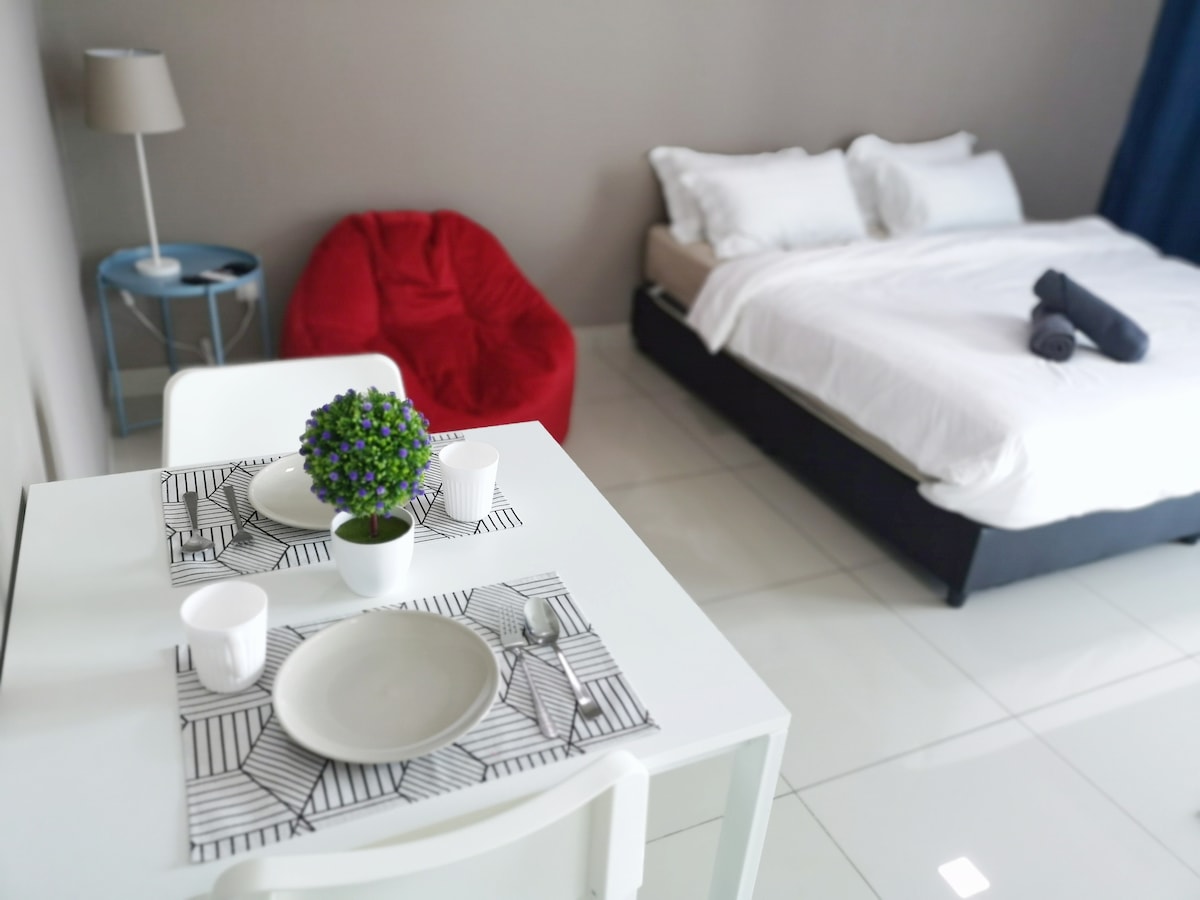 A cozy studio layout features a spacious bed with white linens, complemented by white decorative pillows. A small dining table is set for two, adorned with plates and cups. A red bean bag chair and a bedside lamp add color and warmth to the inviting space.