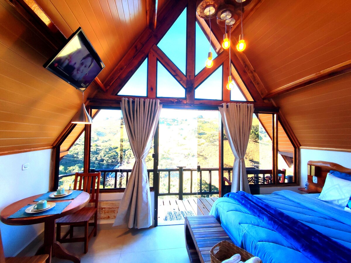 A bright interior showcases a spacious room with large windows offering views of the surrounding mountains. Exposed wooden beams and pendant lighting enhance the charm. A blue bedspread covers the bed, and a small table is set with two plates, suggesting a cozy dining option.