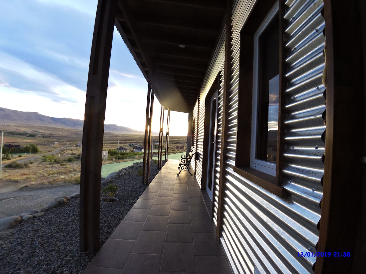 A covered walkway extends alongside a modern structure, featuring corrugated metal siding. The gray tiled floor leads towards scenic views of the surrounding landscape. Natural light casts soft shadows, creating a calming atmosphere. Two benches are positioned along the pathway, inviting relaxation.