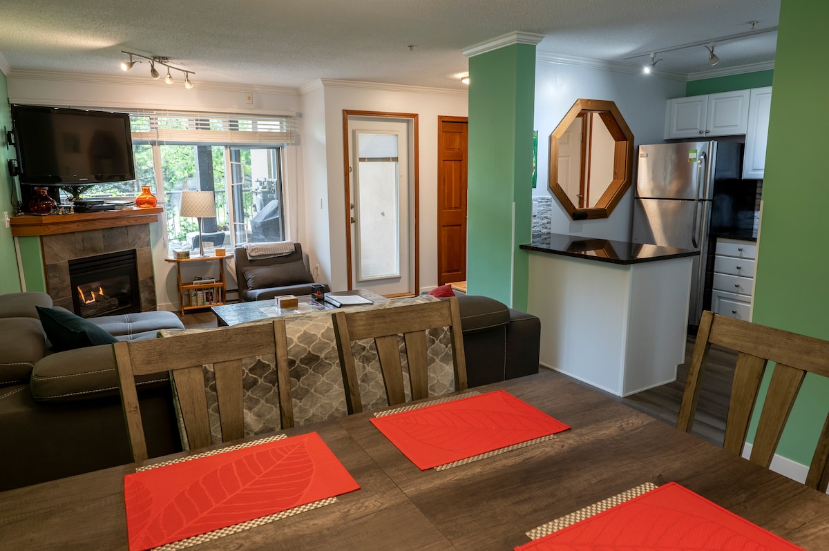 An open living space is showcased with a dining table set for meals, featuring red placemats. A cozy seating area includes a dark sectional sofa near a fireplace. Natural light filters through large windows, while a modern kitchen with stainless steel appliances and granite counters is visible in the background.