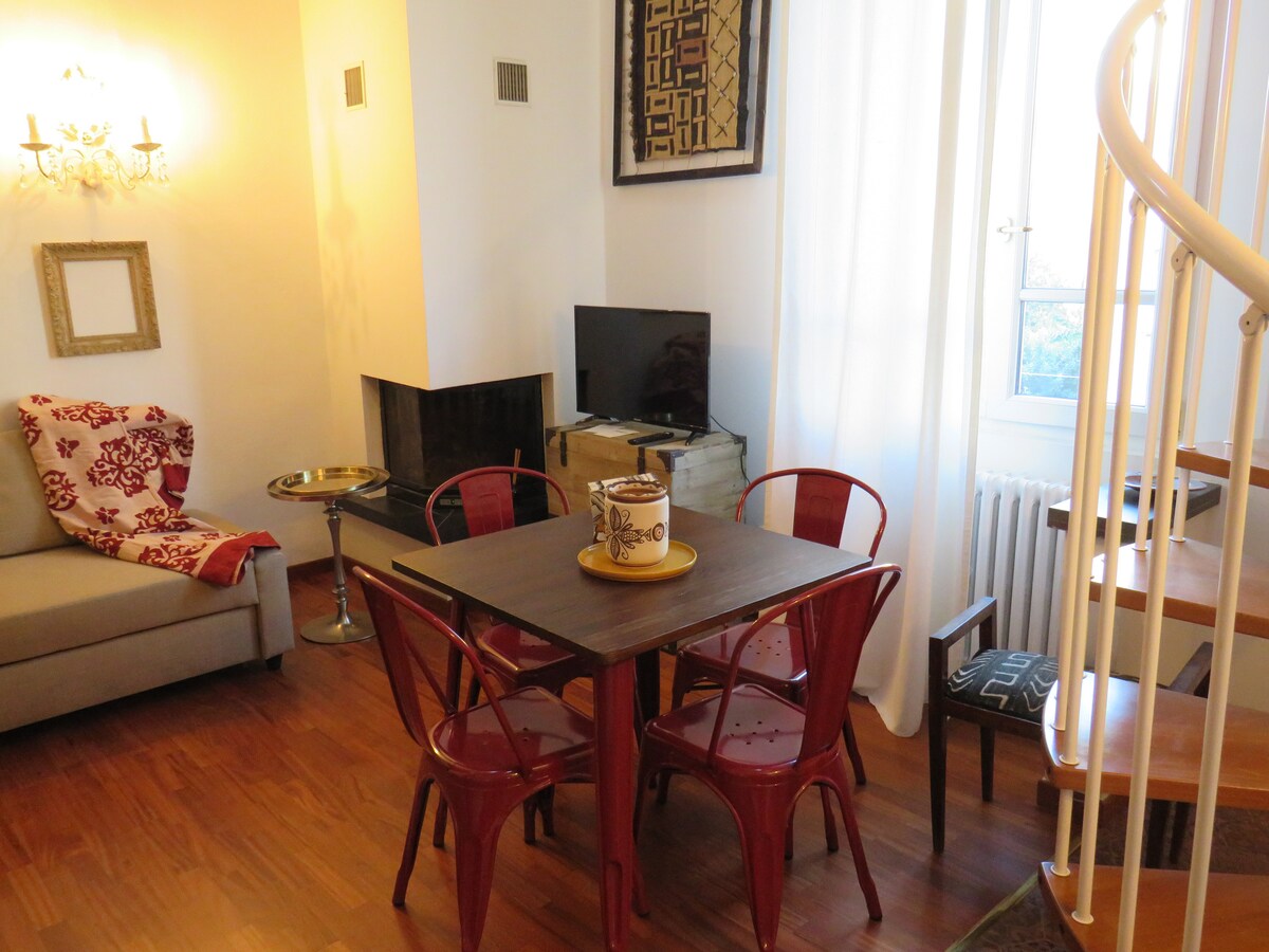 A cozy living area is depicted, featuring a small wooden table surrounded by four red chairs. A comfortable sofa is situated nearby, with a decorative blanket draped over it. A television is placed on a stand across from the seating area, and a spiral staircase is visible in the background.