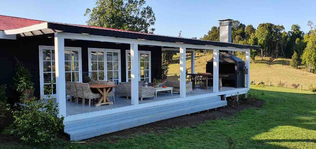 A covered outdoor patio area is furnished with a round wooden table and comfortable seating arrangements. The backdrop features a lush green landscape and a barbecue area, creating an inviting space for relaxation and outdoor dining.