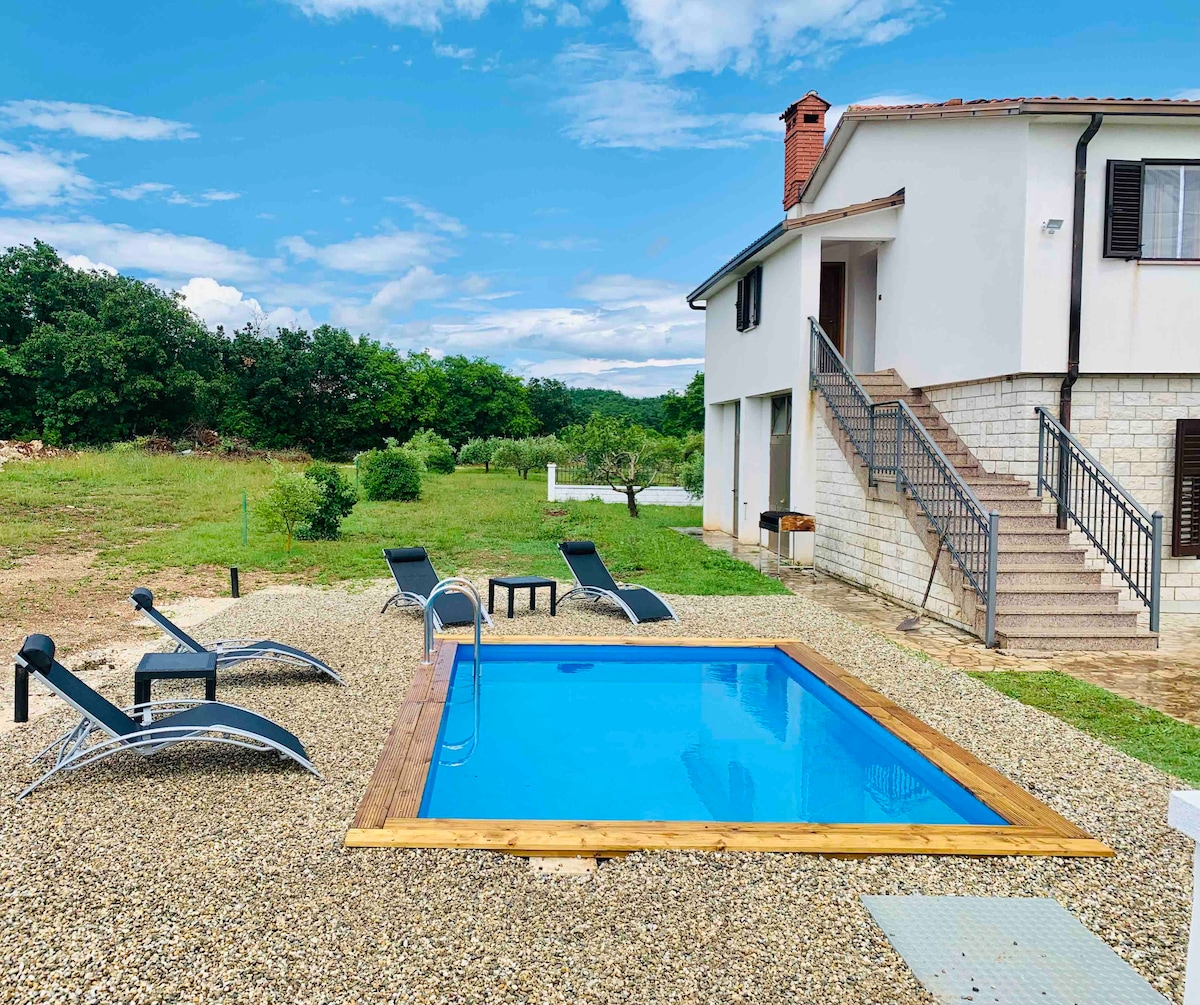 A private outdoor area features a rectangular pool surrounded by wooden decking and gravel. Several lounge chairs are positioned near the pool, and the two-story building stands nearby, complemented by greenery in the background under a clear blue sky.