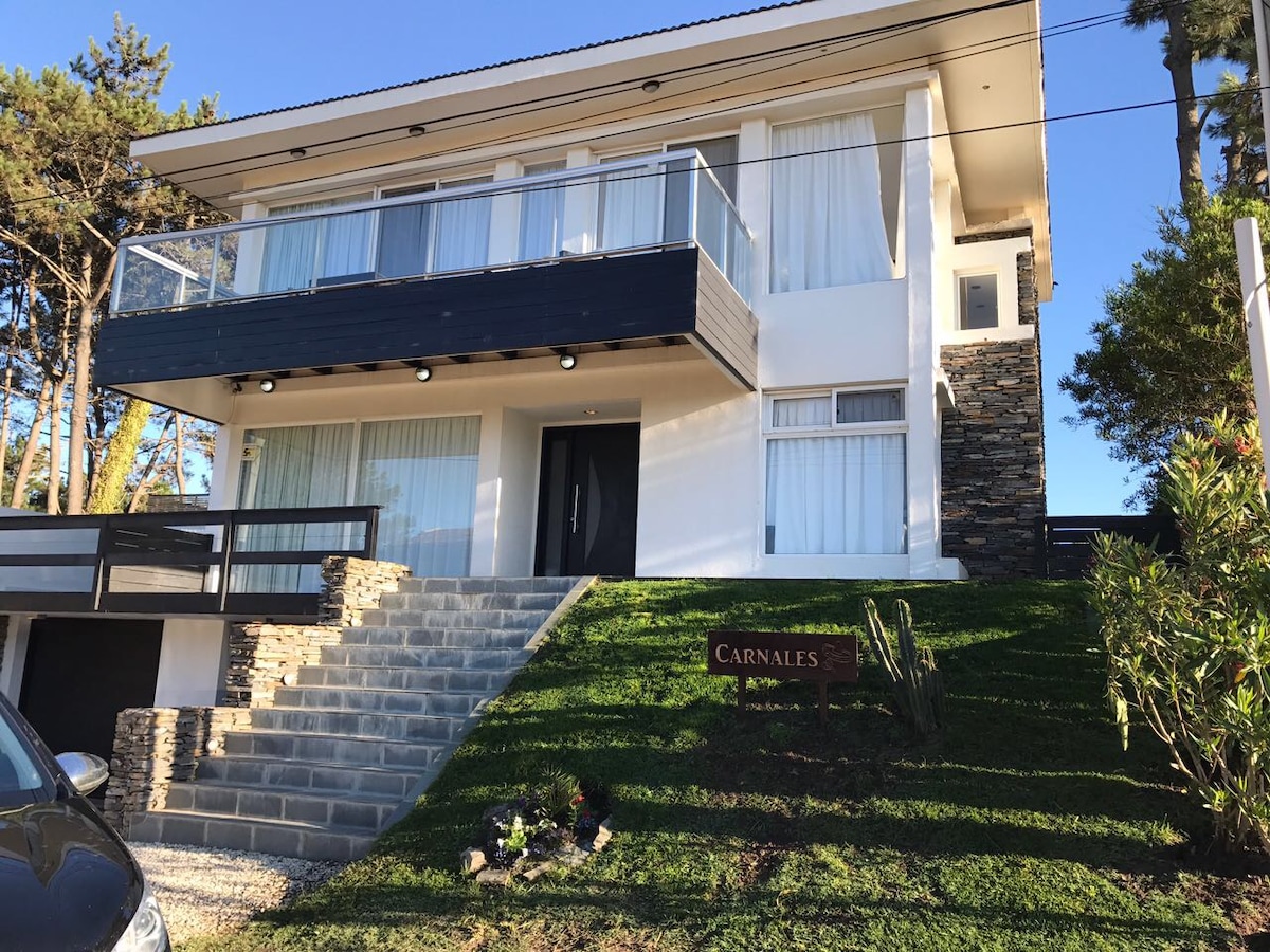 A modern two-story house features large glass windows and a prominent balcony. A well-maintained lawn with decorative plants leads up the stone steps to the entrance. The exterior is a blend of sleek white walls and natural stone accents.