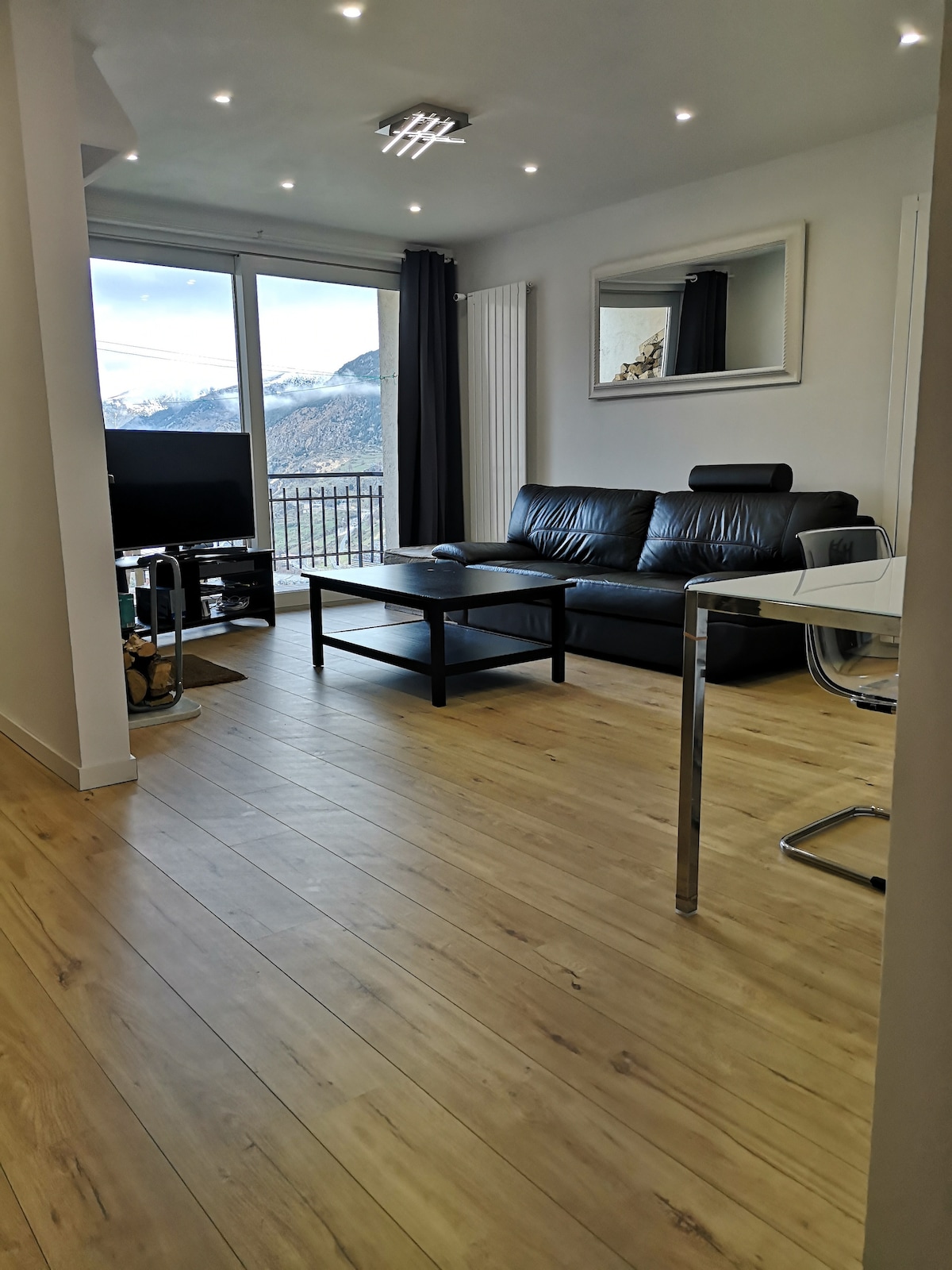 A modern living space features a black leather sofa and a glass coffee table, complemented by a large window that offers mountain views. A television is positioned nearby, while light hardwood flooring and recessed lighting enhance the room's bright and airy feel.