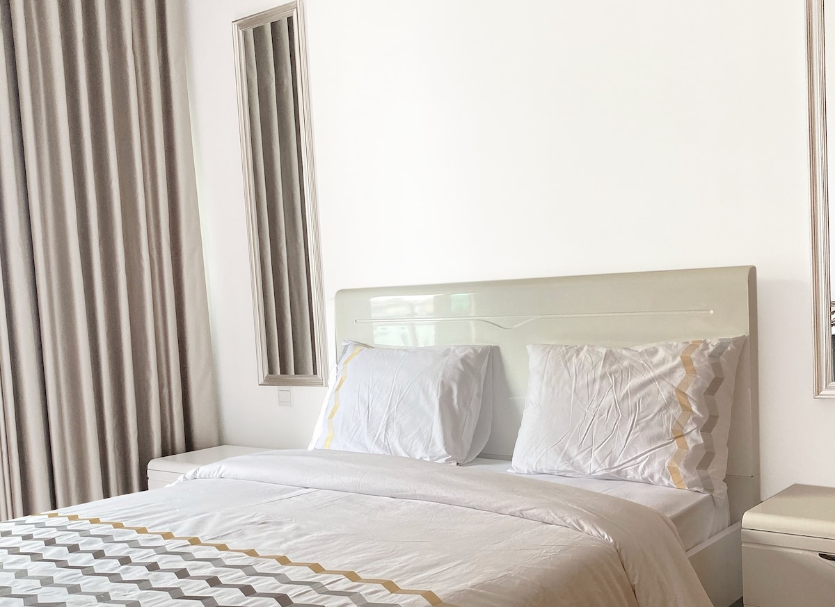 A clean, well-lit sleeping area features a spacious bed with white linens and subtle golden accents. The light-colored walls are complemented by large, soft-textured curtains. Two mirrored panels reflect the natural light coming into the room, enhancing the overall brightness.
