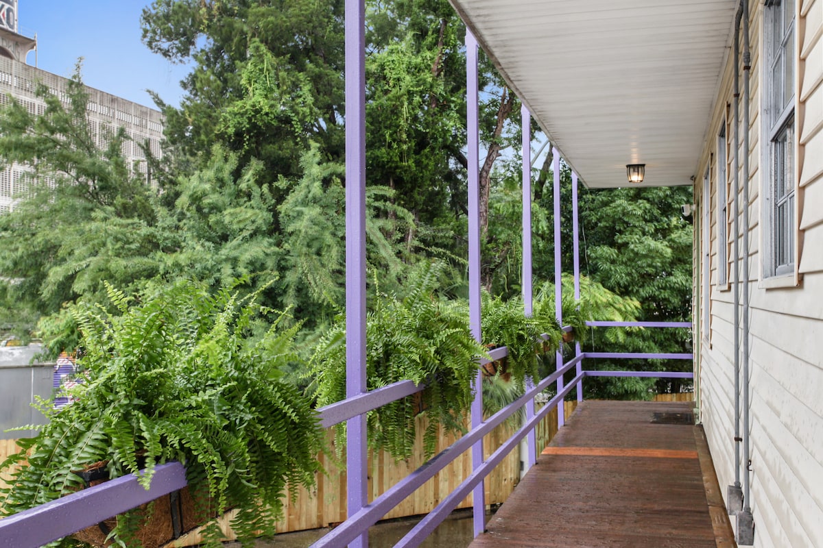 A shaded porch extends along the exterior of the unit, supported by purple railings. Lush ferns are arranged in planters along the edges, providing a vibrant green contrast to the wood flooring. The surrounding trees offer a natural backdrop, enhancing the outdoor experience.