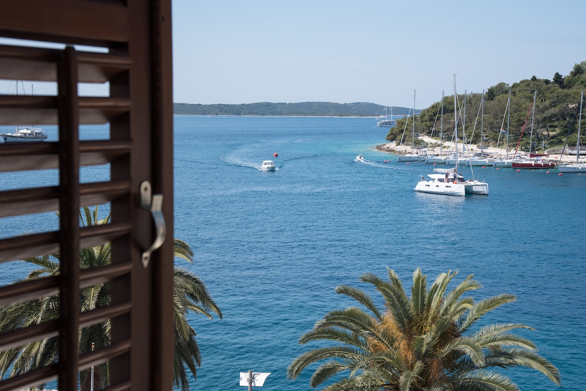 A view of a serene blue sea is framed by partially opened wooden shutters. Palm trees are visible in the foreground, while small boats navigate the water. A coastal landscape with green hills and a marina can be seen in the distance, completing the scene.