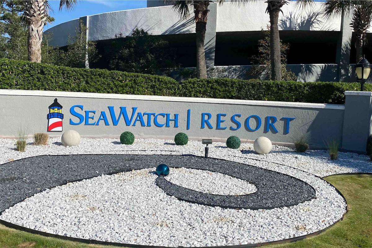 The entrance to SeaWatch Resort is marked by a prominent sign on a landscaped area with decorative white stones, green shrubs, and stylized circular features. Palm trees are visible in the background, contributing to the tropical ambiance of the location.