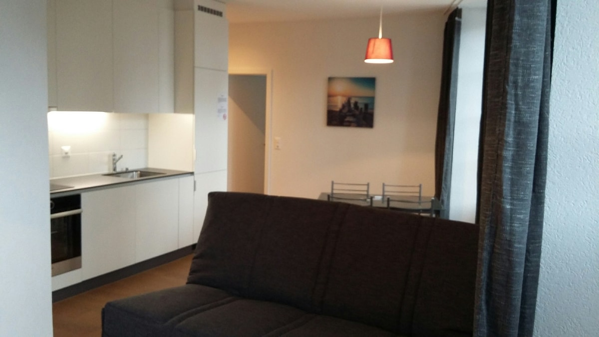 A modern living area is featured, showcasing a dark sofa and a small dining table with metal chairs. The kitchen area is seen in the background, equipped with sleek cabinetry and under-cabinet lighting. A pendant light adds a subtle glow, and a scenic artwork is displayed on the wall.