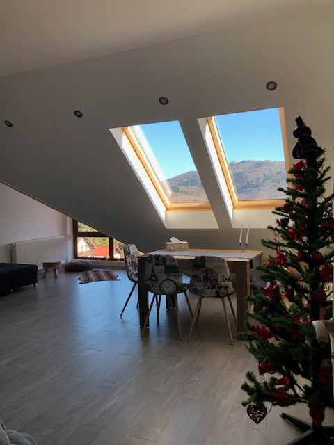 A bright living space is enhanced by large skylights that offer views of the surrounding mountains. A dining table with decorative chairs is positioned beside a Christmas tree. Light wood flooring contributes to a modern and airy feel in the room.