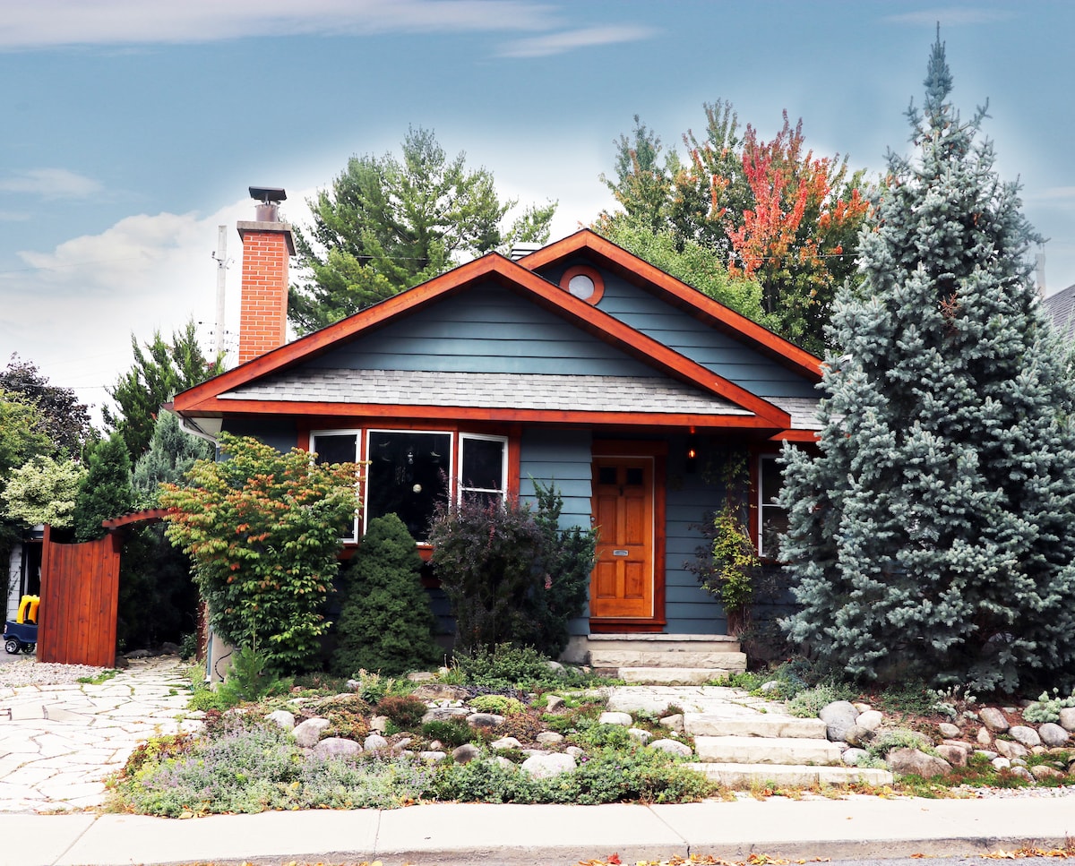 This charming 1939 bungalow features a distinctive blue exterior with red accents. Lush greenery surrounds the entrance, complemented by a stone pathway leading to a wooden front door. The landscaped garden showcases various plants and trees, creating a serene and inviting atmosphere.