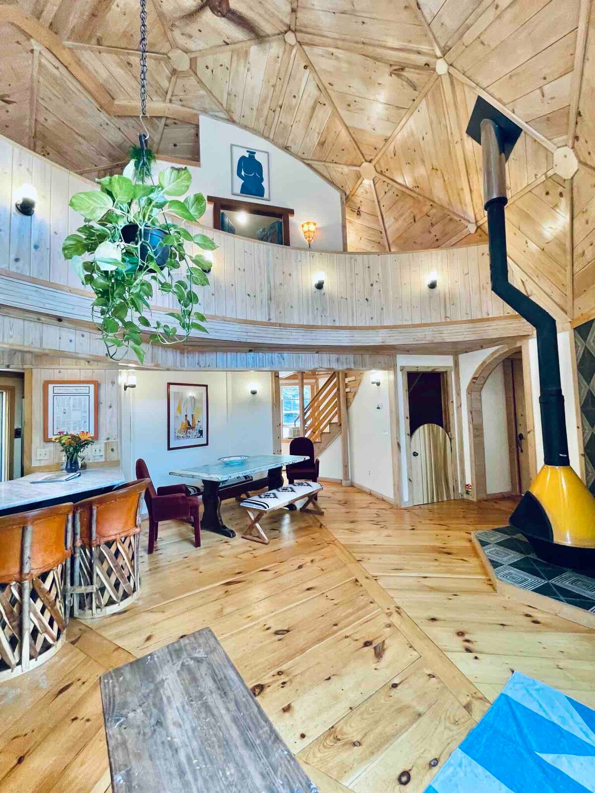 An interior view of a dome home is presented, showcasing natural wood paneling and a spacious layout. A unique yellow wood stove is located near the center, complemented by a cozy seating area and a circular bar with wooden stools. Ample light floods the space from various windows.
