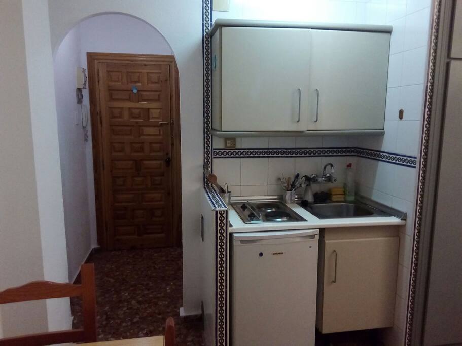 Cocina con nevera, placas eléctricas, fregadero y armario con enseres de cocina.