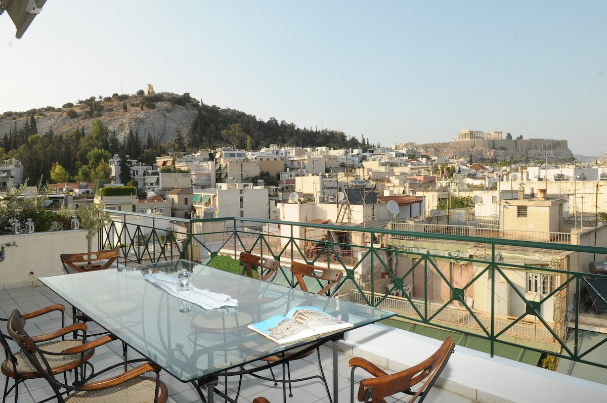 A spacious outdoor terrace is displayed, furnished with a glass table and several wooden chairs. The captivating view includes rooftops stretching across the city, with the Acropolis prominent in the background and the Lycabettus Hill visible to the left.